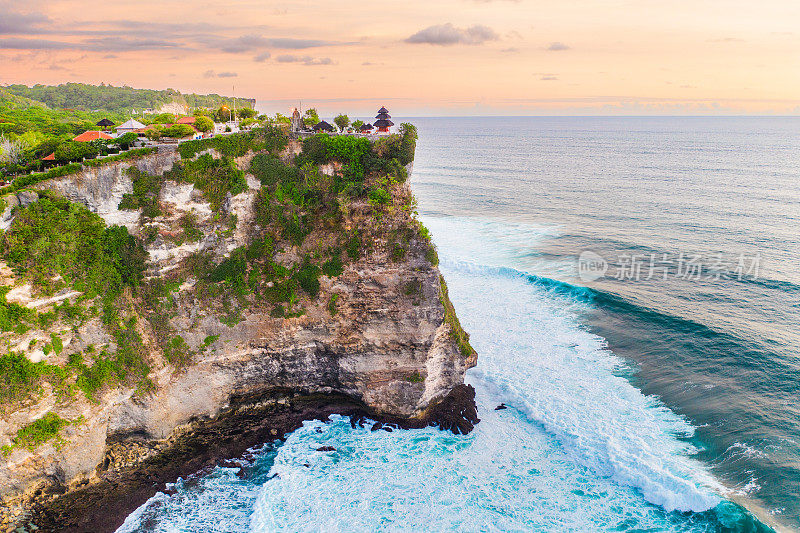 巴厘岛-在日落和乌鲁瓦图神庙(Pura Luhur Uluwatu)中，海浪在蓝色海洋的绿色悬崖上撞击的鸟瞰图。